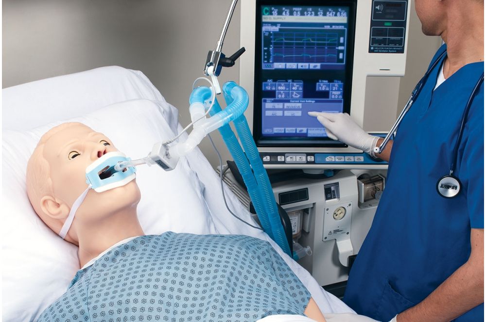 A medical manikin on a hospital bed with an endotracheal tube connected to a ventilator on a nearby medical cart.
