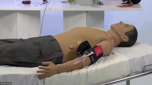 Medical training manikin on an exam table, fitted with a blood pressure cuff and ECG leads on the chest and arms.